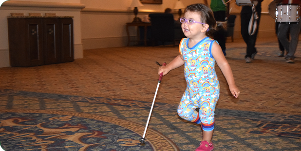 A toddler skips along a hallway with her cane.