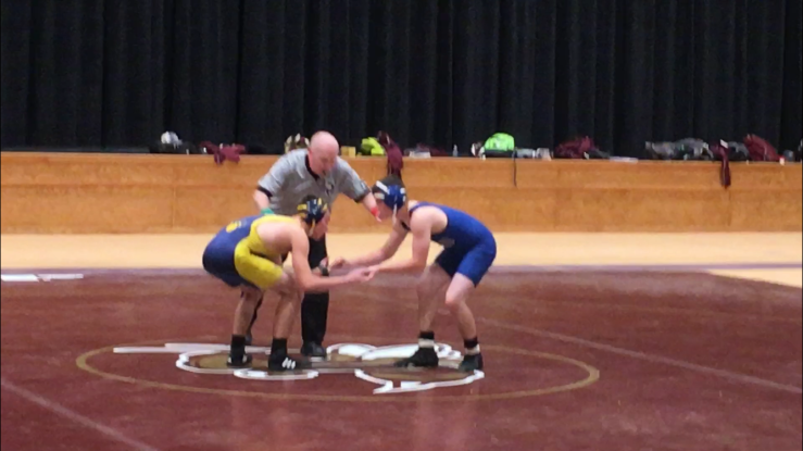 Inside a gym, Matthew and his wrestling opponent face off as the referee prepares to signal the start of the match.